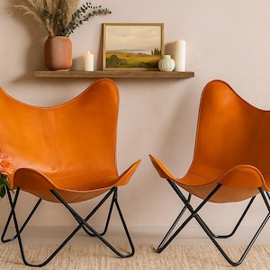 Op de afbeelding: Twee oranje leren vlinderstoelen met zwarte metalen frames. Boven een plank met een ingelijst landschapsschilderij, kaarsen en een vaas. Een vaas met rozen staat voor de stoelen.