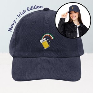 May include: Navy blue corduroy baseball cap with an embroidered design of a beer mug and rainbow. The text "Navy - Irish Edition" is printed on the side of the cap. A woman is wearing a similar cap in a circular inset.