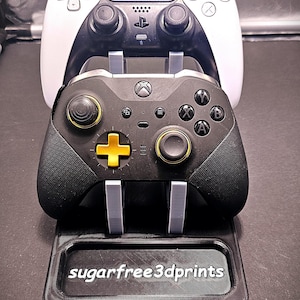 May include: A black and white PlayStation 5 controller and a black Xbox controller with gold accents are displayed on a black 3D printed stand. The stand has the text "sugarfree3dprints" printed on it.