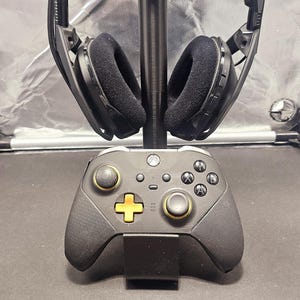 May include: A black gaming headset with a black and gold controller on a stand. The headset has a black headband and earcups with black padding. The controller has a black body with gold buttons and triggers.