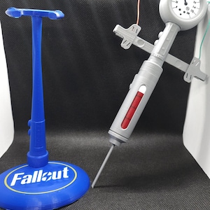 May include: A gray and red Fallout Pip-Boy syringe with a gauge on the top, sitting on a blue stand with the word "Fallout" on it.