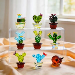 May include: A collection of colorful glass cactus figurines displayed on a clear acrylic shelf. The cacti come in various shapes and colors, including green, blue, and yellow, and are potted in small, decorative pots. Some have butterfly accents.