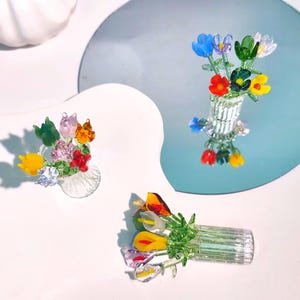 Op de afbeelding: Drie glazen bloemboeketten met kleurrijke bloemen in kleine vazen. De bloemen bevatten gele, rode, blauwe, paarse en groene bloemblaadjes. De vazen zijn van helder glas en de boeketten zijn gerangschikt op een wit oppervlak en een reflecterend oppervlak.