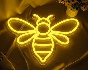 May include: A yellow neon bee sign with a black and yellow striped abdomen. The bee is lit up and glowing against a yellow fabric background.