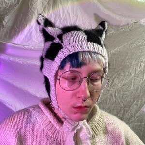 Puede incluir: Una persona lleva una gorra de punto blanca y negra con orejas de gato. La gorra tiene un patrón de cuadros blancos y negros. La persona también lleva un suéter rosa claro y gafas redondas.