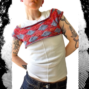 May include: A cropped, hand-knitted shrug featuring a vibrant diamond pattern in shades of red and light blue. The garment has short sleeves and a wide, open neckline, layered over a plain white ribbed tank top and dark blue denim jeans.