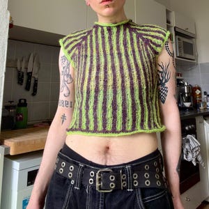 May include: A sleeveless, cropped top with vertical stripes in lime green and purple. The knitted garment has a high neckline and a textured appearance. The top is paired with a black belt and dark-colored pants.