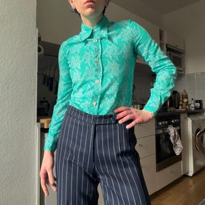 May include: A teal green button-down shirt with a textured pattern is paired with navy blue pinstripe trousers. The shirt has long sleeves and a classic collar. The trousers have a high waist and a straight leg.