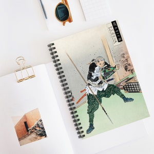 Könnte beinhalten: Spiralgebundenes Notizbuch mit einem japanischen Holzschnitt eines Samurai-Kriegers in Aktion. Das Kunstwerk zeigt eine dynamische Szene mit dem Krieger, der einen Speer und ein Schwert schwingt und traditionelle Kleidung trägt.