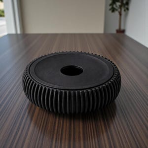 May include: A black, circular container with a ridged, cylindrical exterior and a flat top with a central hole. The container sits on a dark wood surface. The design is modern and minimalist.