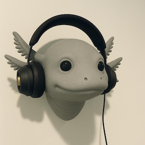 Puede incluir: Una escultura de cabeza de ajolote gris con auriculares negros. El ajolote tiene ojos grandes y oscuros y branquias plumosas. Los auriculares tienen auriculares negros con detalles dorados y una diadema negra. Cuelga un cable negro.