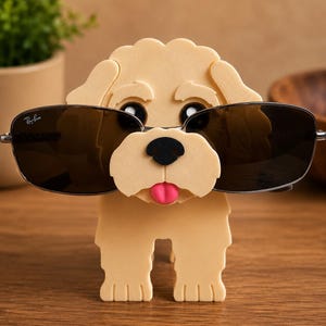 May include: A tan-colored dog-shaped eyeglass holder with black eyes, a black nose, and a pink tongue. The holder is designed to hold a pair of sunglasses with dark lenses and silver frames. The dog has a playful, cartoonish appearance.