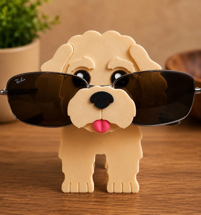 May include: A tan-colored dog-shaped eyeglass holder with black eyes, a black nose, and a pink tongue. The holder is designed to hold a pair of sunglasses with dark lenses and silver frames. The dog has a playful, cartoonish appearance.