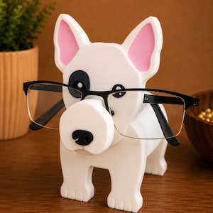 May include: A white dog-shaped eyeglasses holder with pink inner ears and a black eye patch. The holder is wearing black-framed glasses. The dog is standing on a wooden surface.