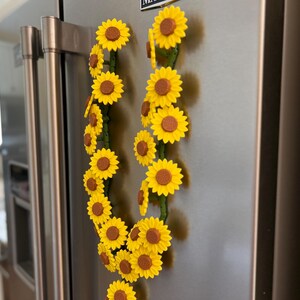 May include: Two decorative sunflower garlands with yellow petals and brown centers are attached to a stainless steel refrigerator. The sunflowers are arranged along green stems, creating a cheerful, summery aesthetic.
