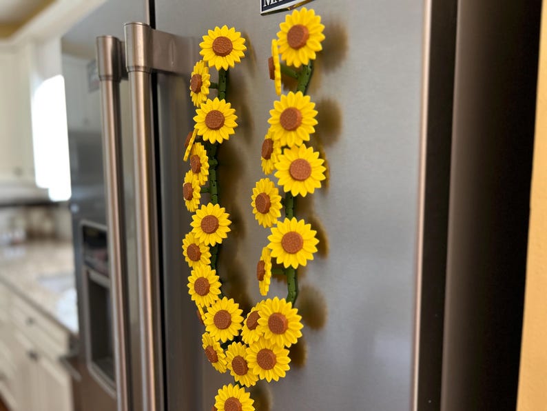 May include: Two decorative sunflower garlands with yellow petals and brown centers are attached to a stainless steel refrigerator. The sunflowers are arranged along green stems, creating a cheerful, summery aesthetic.