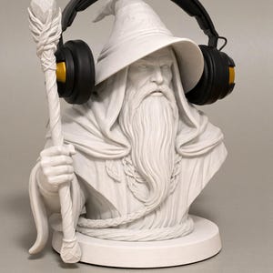 May include: A white bust of a wizard with a long beard, holding a staff, and wearing black headphones with gold accents. The wizard is wearing a hat and a robe. The bust is on a round base.