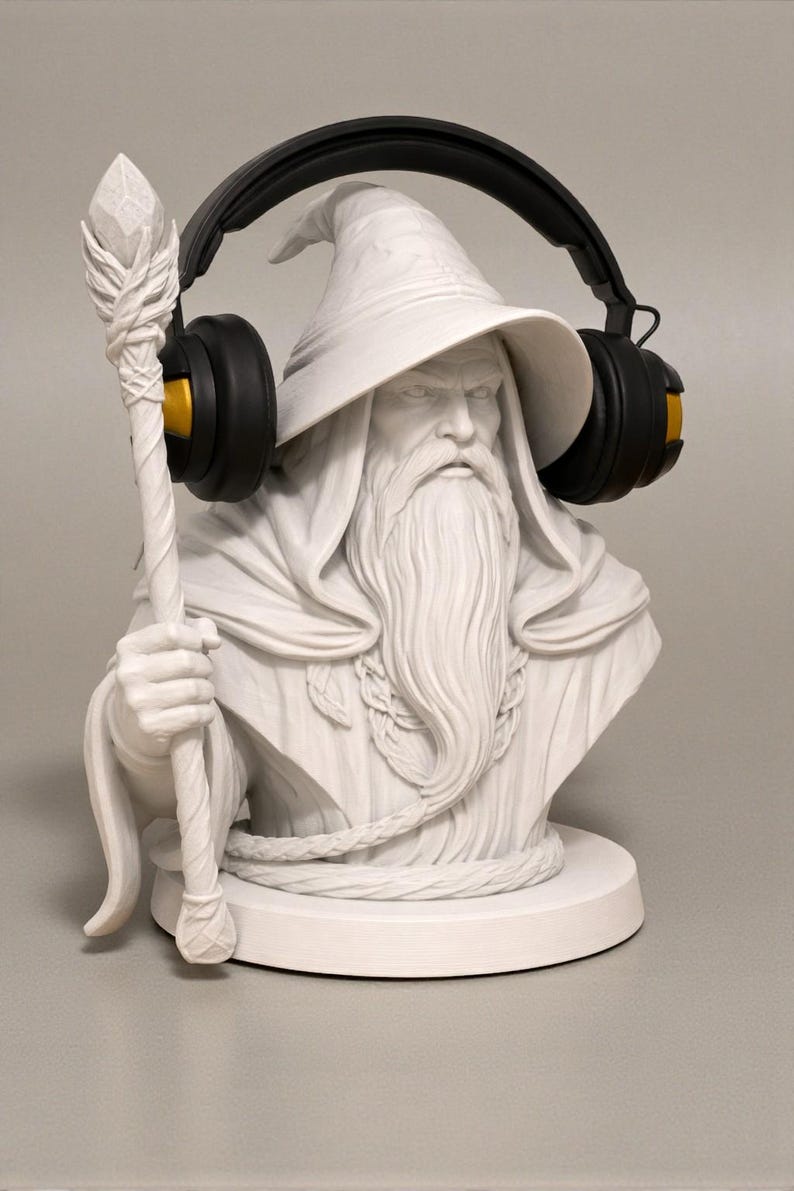 May include: A white bust of a wizard with a long beard, holding a staff, and wearing black headphones with gold accents. The wizard is wearing a hat and a robe. The bust is on a round base.