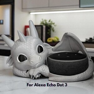 May include: A gray dragon-shaped speaker holder for an Alexa Echo Dot 3. The dragon has large eyes, horns, and wings, cradling the speaker. The text "For Alexa Echo Dot 3" is at the bottom.