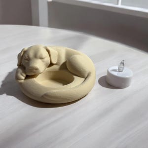 Puede incluir: Un portavelas con forma de perro de color crema con un dise&ntilde;o de perro durmiendo. Junto a &eacute;l, una peque&ntilde;a vela de t&eacute; blanca que funciona con pilas con una llama clara y parpadeante.