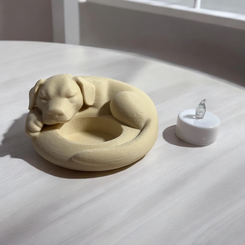 Puede incluir: Un portavelas con forma de perro de color crema con un dise&ntilde;o de perro durmiendo. Junto a &eacute;l, una peque&ntilde;a vela de t&eacute; blanca que funciona con pilas con una llama clara y parpadeante.