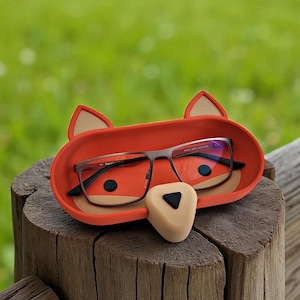May include: A red fox-shaped glasses holder with a beige nose and ears. The holder contains a pair of eyeglasses with dark frames. The holder is resting on a wooden surface, with a green background.