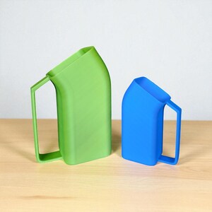 May include: Two modern pitchers, one green and one blue, sit side-by-side on a wooden surface. Each pitcher has a rectangular body with a slanted top and a handle. The green pitcher is larger than the blue one.