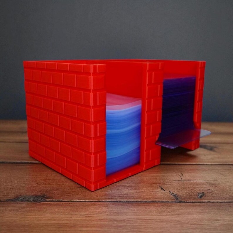 May include: A red card storage box, designed with a brick pattern. The box has a slot for easy access and contains a stack of blue and purple cards, ideal for organising trading cards or other collectibles.
