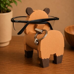 May include: A brown bear-shaped eyeglasses holder with black-framed glasses resting on it. The holder has dark brown ears and feet, and a silver key ring is attached to its back. The holder is made of cardboard and sits on a wooden surface.