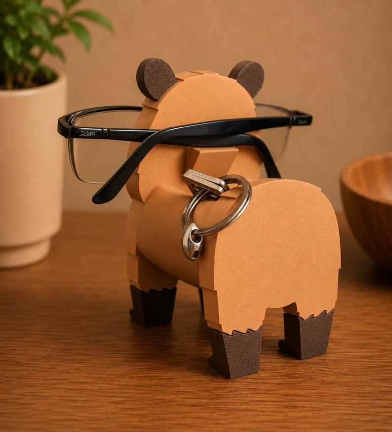 May include: A brown bear-shaped eyeglasses holder with black-framed glasses resting on it. The holder has dark brown ears and feet, and a silver key ring is attached to its back. The holder is made of cardboard and sits on a wooden surface.