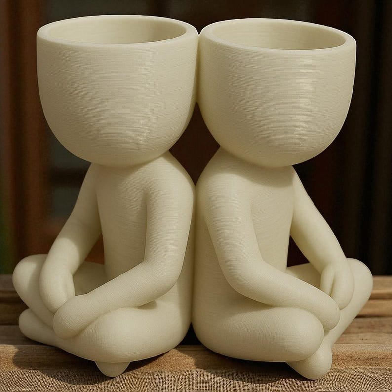 May include: Two off-white ceramic planters shaped like seated figures with open-top heads. The figures are in a meditative pose, with their legs crossed and arms resting on their knees. The planters are suitable for small plants or succulents.