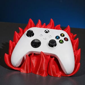Peut inclure: Une manette Xbox blanche repose dans un support rouge en forme de flamme. La manette a des joysticks noirs et des boutons colorés. Le support est d'un rouge vif, créant un contraste saisissant.
