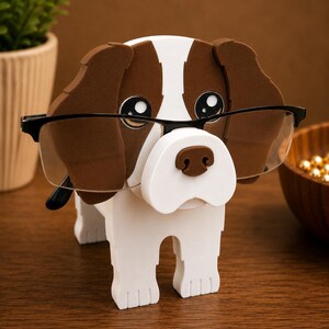 May include: A whimsical dog-shaped eyeglasses holder. The holder is white with brown accents on the ears and nose. It features large, cartoonish eyes and is designed to hold a pair of eyeglasses. The glasses are black-framed.