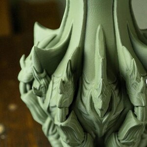 May include: Close-up of a sage green, ornate decorative object with a textured surface. The object features a series of pointed, leaf-like shapes and intricate details, suggesting a sculptural or artistic design. The overall aesthetic is organic and stylized.