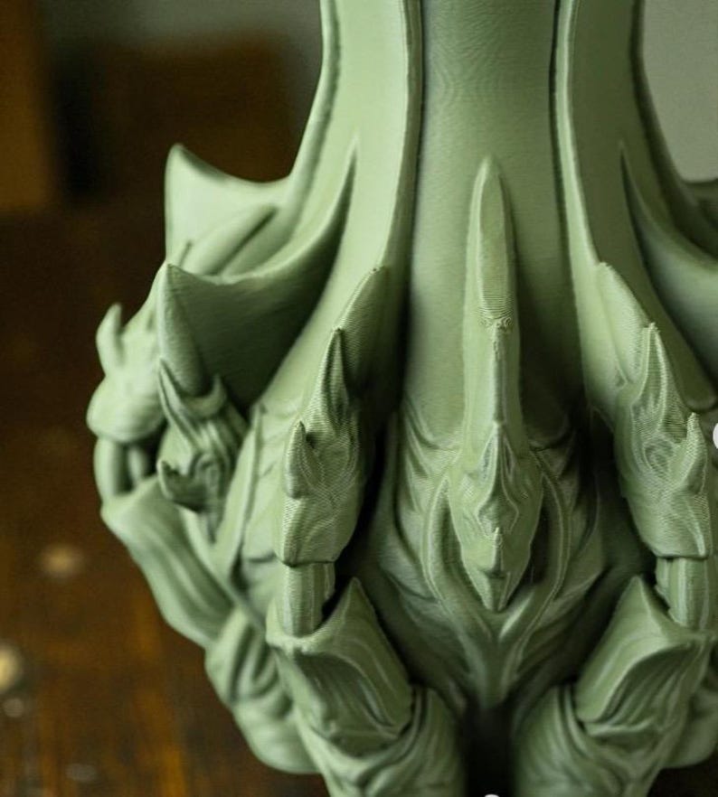 May include: Close-up of a sage green, ornate decorative object with a textured surface. The object features a series of pointed, leaf-like shapes and intricate details, suggesting a sculptural or artistic design. The overall aesthetic is organic and stylized.