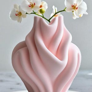 May include: A soft pink vase with a unique, flowing design, holding white orchid flowers. The vase has a sculpted, wavy texture. The orchids have white petals with yellow and red accents.