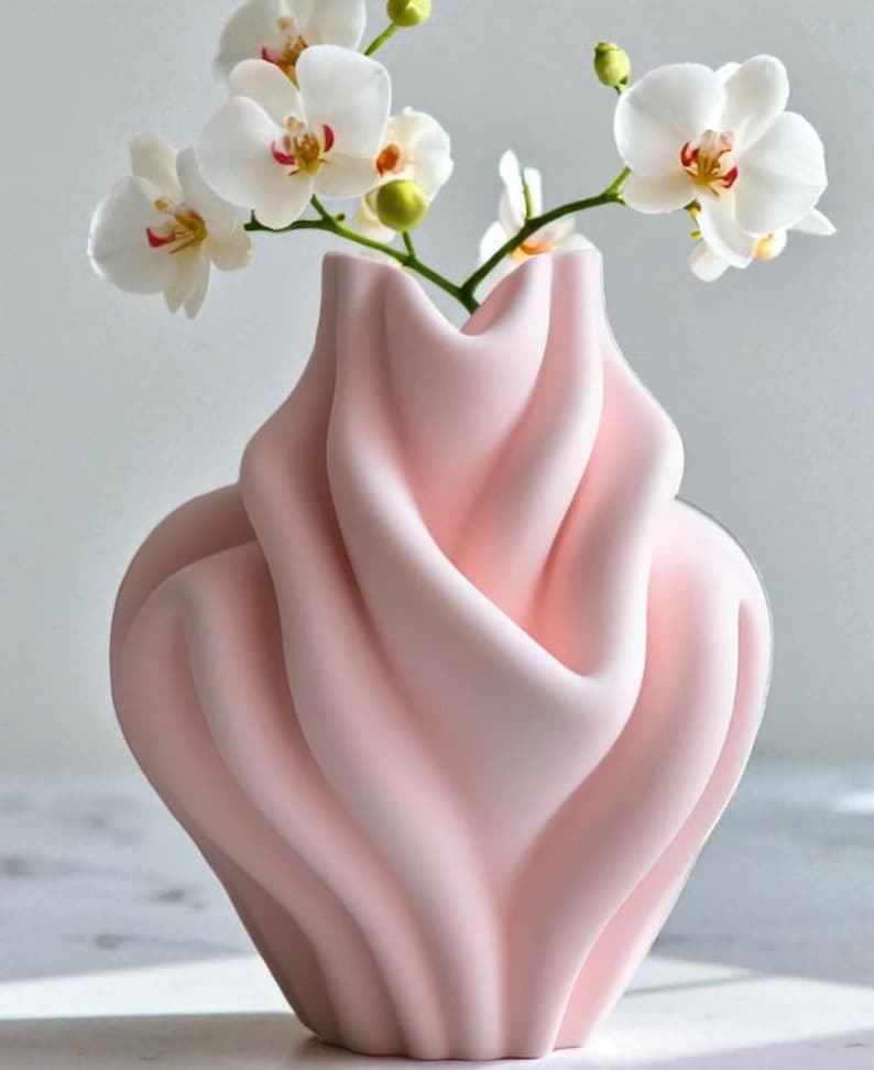 May include: A soft pink vase with a unique, flowing design, holding white orchid flowers. The vase has a sculpted, wavy texture. The orchids have white petals with yellow and red accents.