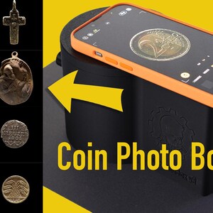 Op de afbeelding: Een zwarte muntfotobox met een oranje rand en de tekst "Coin Photo Box v3" aan de voorkant. Een smartphone ligt op de doos, met een munt zichtbaar op het scherm.
