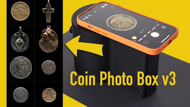 Op de afbeelding: Een zwarte muntfotobox met een oranje rand en de tekst "Coin Photo Box v3" aan de voorkant. Een smartphone ligt op de doos, met een munt zichtbaar op het scherm.