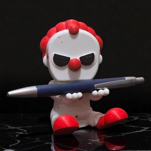 May include: A white and red clown figurine holding a blue pen. The clown has a red nose, red hair, and black eyes. The pen is silver and blue. The figurine is sitting on a black surface.