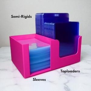 May include: A bright pink card storage organizer with three compartments. The compartments are labeled "Sleeves", "Toploaders", and "Semi-Rigids". The organizer holds blue card sleeves and blue toploaders. The organizer is on a white marble surface.