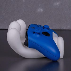 May include: A white, hand-shaped gaming controller holder cradles a blue controller. The holder is made of a smooth, solid material. The controller has black buttons and a black joystick.