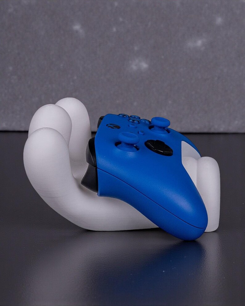 May include: A white, hand-shaped gaming controller holder cradles a blue controller. The holder is made of a smooth, solid material. The controller has black buttons and a black joystick.