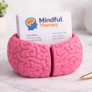 May include: A pink brain-shaped business card holder with the words "Mindful Therapy" printed on the cards inside. The cards also display a logo of a brain and a profile. A gold pen and paperclips are on the table.