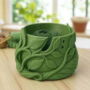 May include: A green yarn bowl with a leaf and vine design. The bowl is filled with green yarn. The bowl has two holes on the side. The bowl is sitting on a wooden surface.