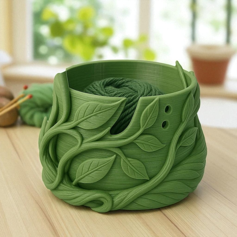 May include: A green yarn bowl with a leaf and vine design. The bowl is filled with green yarn. The bowl has two holes on the side. The bowl is sitting on a wooden surface.