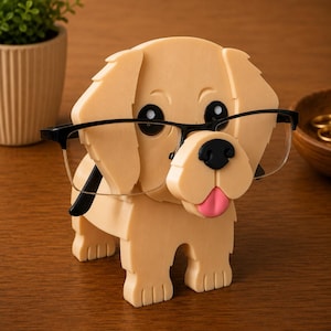 May include: A tan-colored dog-shaped eyeglasses holder with black-framed glasses. The dog has black eyes, a black nose, and a pink tongue. The holder is made of a smooth material and sits on a wooden surface.