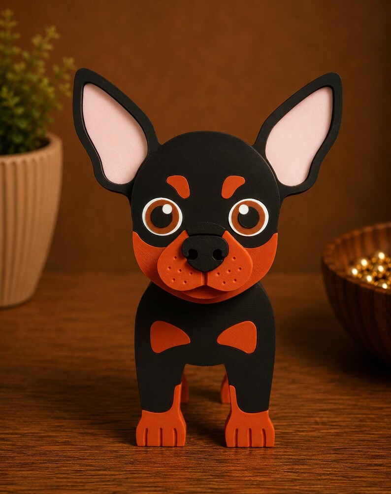 May include: A black and orange dog figurine with large ears and expressive eyes. The dog has a black body with orange accents on its face, paws, and chest. The ears are black with pink interiors. The figurine is standing on a wooden surface.