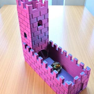 May include: A pink and purple dice tower and tray set, designed to resemble a castle. The tower has a rectangular shape with crenellations and small windows. A multi-sided die rests in the tray. The set is made of plastic.