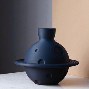 Może przedstawiać: Granatowy ceramiczny wazon o unikalnym planetarnym designie. Wazon ma kulistą podstawę z pierścieniem i cylindryczną g&oacute;rą. Powierzchnia ma kilka okrągłych wgłębień, przypominających kratery.